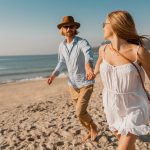 young-attractive-smiling-happy-man-hat-blond-woman-white-dress-running-together-beach-min