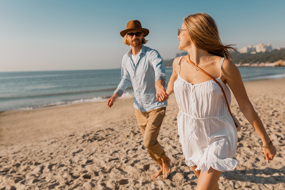 young-attractive-smiling-happy-man-hat-blond-woman-white-dress-running-together-beach-min